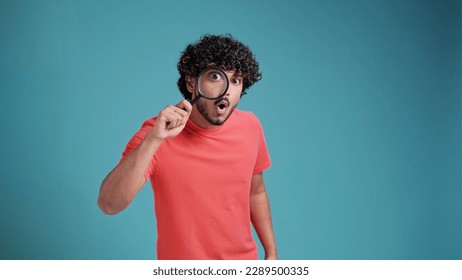 Funny man looking through magnifying glass, searching or investigating something, standing in orange sweater against turquoise background