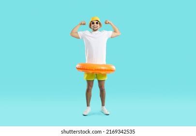 Funny man with orange inflatable swimming circle flaunts his strength on light blue background. Full length of playful strong male swimmer in beachwear showing off his biceps. Summer vacation concept.