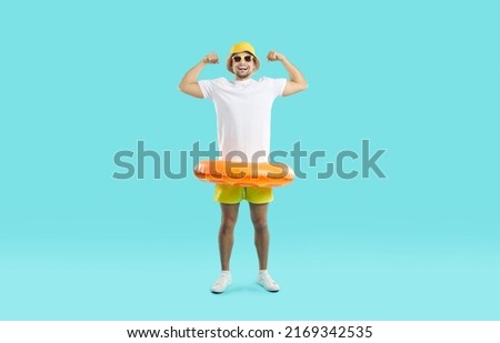 Funny man with orange inflatable swimming circle flaunts his strength on light blue background. Full length of playful strong male swimmer in beachwear showing off his biceps. Summer vacation concept.