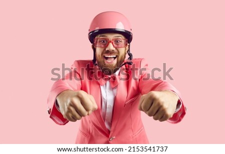 Funny man in pink protective helmet pretends to be motorcycle rider isolated on pink background. Cheerful man in pink formal in suit, glasses and helmet with crazy expression looks at camera. Banner.