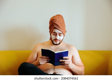 Funny Man Reading Book Sitting On Sofa Relaxing At Home, Pamper Wellness Caucasian Male Pampering Skin Care With Facial Mask Treatment