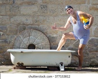Funny man in retro swimsuit going to the bathtub outdoor. Swimmer with a bathing cap goes into the bath with toy inflatable duck. Cheerful man goes to relax in the tub.