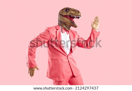 Funny man in rubber dinosaur mask dancing and having fun in the studio. Happy lizard headed guy in stylish funky vibrant pink party suit doing Egyptian dance moves isolated on pink colour background