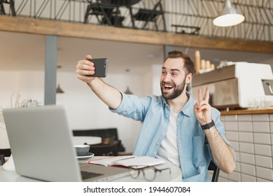 Funny man sit alone at table in coffee shop cafe restaurant indoors working studying on laptop computer doing selfie shot on cell phone showing victory sign. Freelance mobile office business concept - Shutterstock ID 1944607084