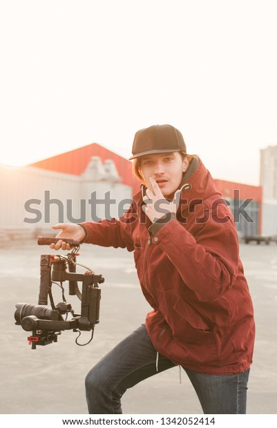Funny man with a stabilizer and\
camera on the background of the sunset. Portrait of a young\
videographer holding a stabilizer in his hands and posing on the\
camera.