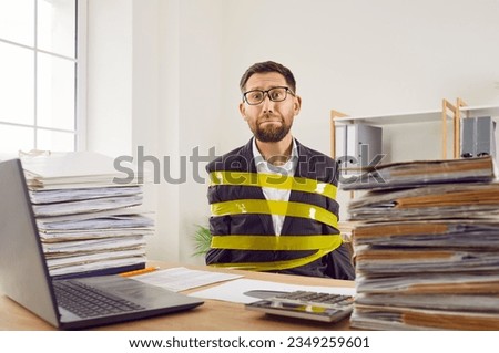 Funny man, stuck in office like work slave, bound with yellow adhesive tape sitting at desk with notebook computer and piles of paper folders and binders and looking at camera with sad face expression