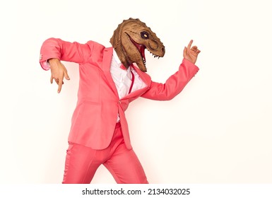 Funny man wearing a wacky dinosaur mask having fun at a crazy party. Profile view of a happy young guy in a pink suit and a silly scary ugly masquerade dino mask dancing isolated on a white background