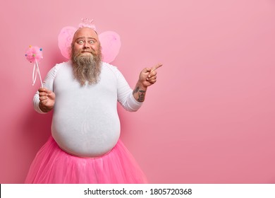 Funny man wears fairy costume, invites you on holiday or costume party, indicates right at blank space, holds magic wand, poses against rosy wall. Dad entertains children during birthday celebration - Shutterstock ID 1805720368