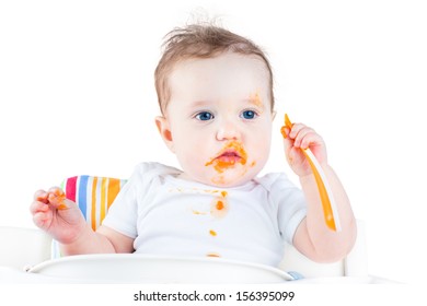 Funny Messy Baby Eating Her First Solid Vegetable Food In A White High Chair, Isolated On White