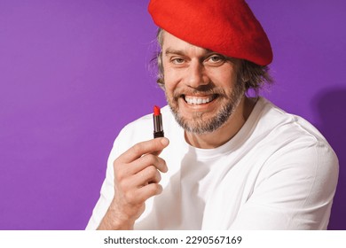 Funny middle aged man wearing red beret is holding lipstick in his hand against purple background.
