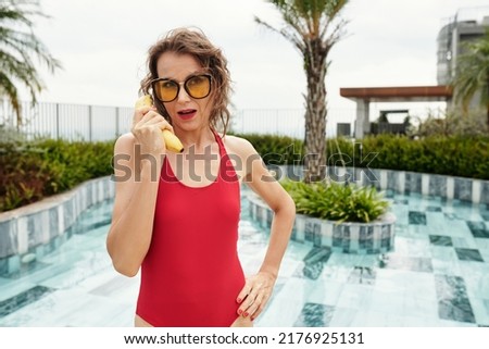 Funny middle-aged woman in yellow glasses and red swimsuit pretending to make call with banana