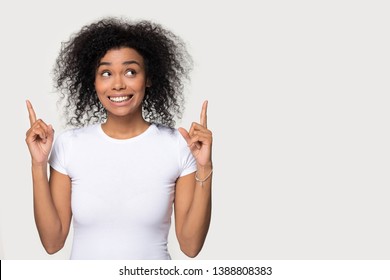 Funny mixed race woman in white t-shirt curly afro hairs pointing fingers and looking up standing aside over grey blank wall advertise product beauty salon professional care copyspace for text concept