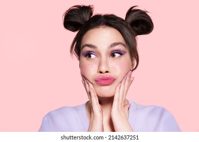 Funny modern beautiful young girl with trendy lavender make up, pretty hairdo of two hair buns looking aside, touching cheeks, pouted lips. Stylish pensive teen with lilac eyeshadow feel puzzlement.