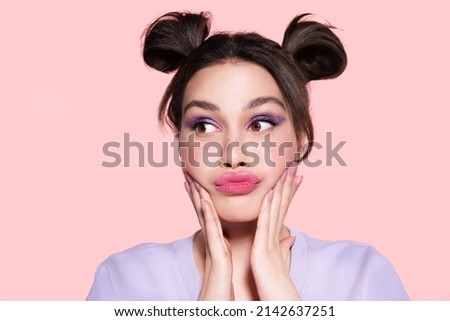 Funny modern beautiful young girl with trendy lavender make up, pretty hairdo of two hair buns looking aside, touching cheeks, pouted lips. Stylish pensive teen with lilac eyeshadow feel puzzlement.