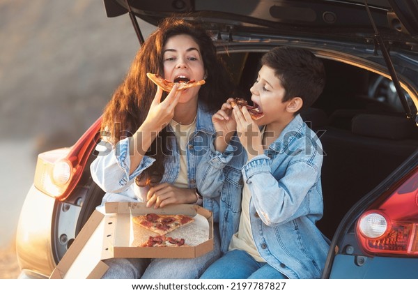Funny mom with teen son sitting in the trunk of\
a car and eating slices of take away pizza from box. Happy mother\'s\
day concept.