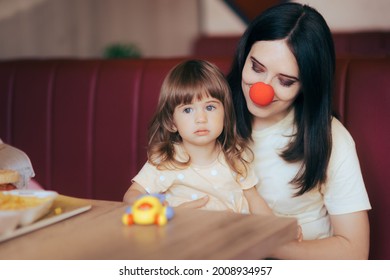 Funny Mom Wearing Clown Nose Celebrating Daughter In A Restaurant. Mother Entertaining Her Toddler Child For Its Birthday Throwing A Party In A Fast Food Diner
