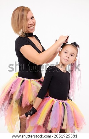 Funny Mother and daughter in same outfits posing on studio weared tutu skirts on the white background. Mom makes a hairstyle for her little girl
