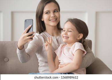Funny mother holds smartphone take selfie with small daughter showing V peace sign, family younger older sister spending free time using electronic gadget having fun with wireless modern tech concept
