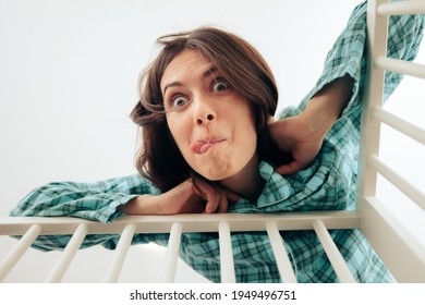 Funny Mother Making Faces Cheering Up Baby In The Crib. Goofy Mom Trying To Calm Her Baby Crying And Suffering. Parent In Nursery Room Comforting Newborn
