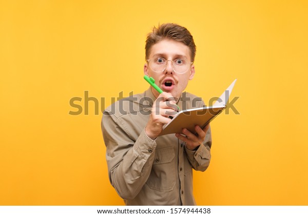 Funny nerd in glasses\
and shirt looks surprised at the camera and writes in a notebook on\
a yellow background. Portrait of funny student writes in exercise\
book, isolated.