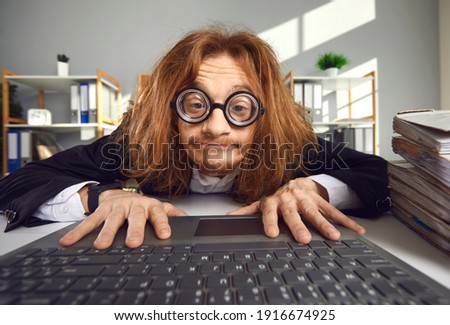 Funny nerd in round thick lens glasses sitting at desk and using laptop. Crazy looking office worker or computer geek typing on keyboard, searching for information on the Internet or doing accounts
