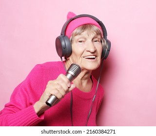 Funny old lady listening music with headphones and singing with mic over pink background