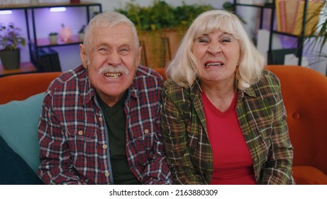 Funny Old Senior Elderly Family Couple Grandparents Man Woman Making Playful Silly Facial Expressions And Grimacing, Fooling Around, Showing Tongue. Mature Grandmother Grandfather On Couch At Home