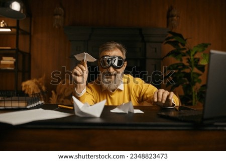 Funny overjoyed mature man wearing pilot goggles playing with origami plane