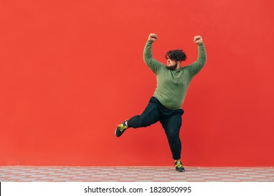 Funny Overweight Dancer Listens To Music On Headphones And Dances On A Red Background, Looking Away At Copy Space. The Fat Man Shows A Dance Performance On A Background Of A Red Wall.