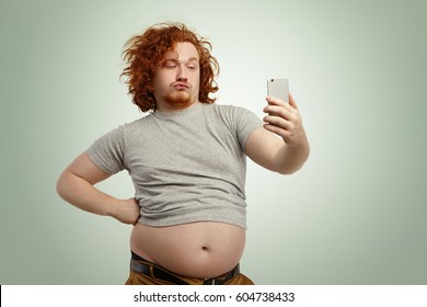 Funny overweight plump man with duck lips wearing undersize t-shirt with belly hanging out of pants, keeping hand on waist, posing for selfie, holding cell phone, trying to seem attractive and sexy