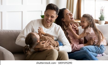 Funny Parents Play With Two Daughters Resting On Couch In Light Cozy Living Room. Family Enjoy Playtime Together At Home, Tickling Each Other Laughing Feels Happy. Having Fun At Home With Kids Concept