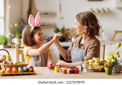 Funny Photo Of Joyful Happy Family Mom And Little Daughter Having Fun And Fooling Around While Decorating Easter Eggs Together For Spring Holidays, Sitting At Table In Cozy Kitchen.  