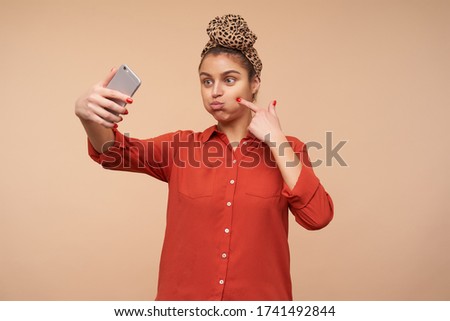 Funny photo of young cheerful brown haired lady puffing out her cheeks while fooling and making selfie with her smartphone, isolated over beige background
