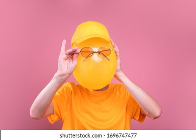 Funny picture of incognito guy hiding face behind yellow balloon. Hold sunglasses in front of it. Guy wear yellow cap and shirt. Isolated over pink background
