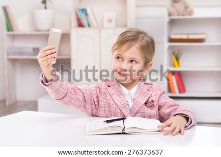 Funny picture of little cute girl playing role of business woman. Girl wearing pink suit. Girl sitting at table with notebook and making photo by phone. Office interior as a background