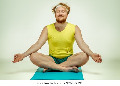 Funny picture of red haired, bearded, plump man on white background. Man wearing sportswear. Man sitting on the mat, he is meditating