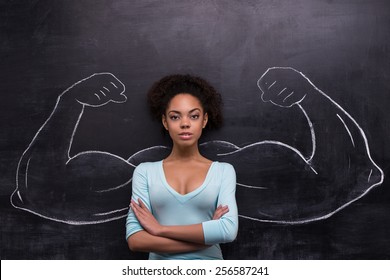 Funny picture of young afro-american woman on chalkboard background seriously looking at camera. Two strong muscular arms painted on chalkboard