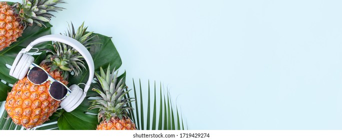 Funny pineapple wearing white headphone, concept of listening music, isolated on blue background with tropical palm leaves, top view, flat lay design.