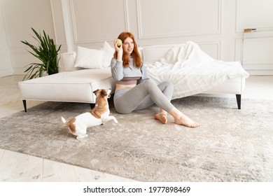 Funny Play With Ball. Ginger Haired Beautiful Young Girl Throws Ball To Small Active Jumping Dog Jack Russell Terrier. Funny Weekend Time At Home. Spacious Living Room With White Sofa And Carpet