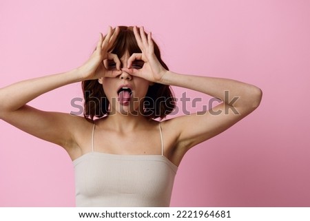 a funny, playful woman with short hair poses standing on a pink background showing the OK sign with her fingers near her eyes, covering them with her hands. Horizontal photo with empty space 