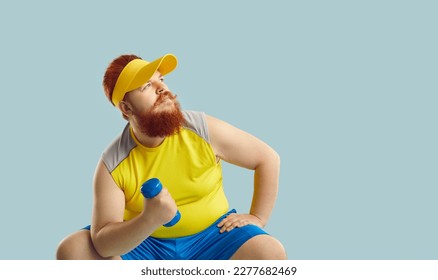 Funny plump fat man with ginger beard, in yellow top sitting on blue studio background, holding workout dumbbell, training muscles, thinking and looking at blank text copyspace for inspiring quote - Powered by Shutterstock