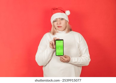 Funny plus size woman portrait in white sweater and santa hat on red background, holding smartphone with green screen, chroma key, copy space - Powered by Shutterstock