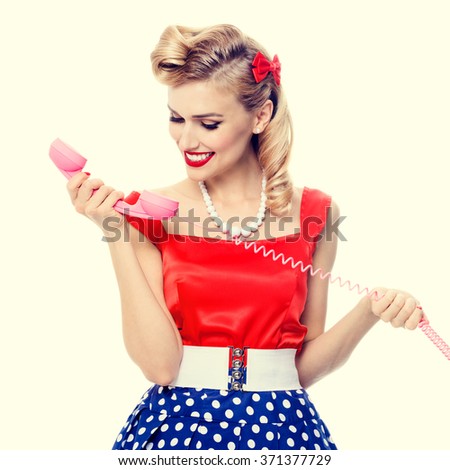Funny portrait of beautiful young happy smiling woman with phone, dressed in pin-up style dress in polka dot. Caucasian blond model posing in retro fashion and vintage concept studio shoot.