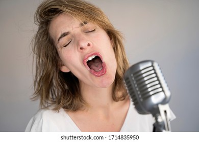 Funny Portrait Of Caucasian Woman Singing A Song With A Vintage Silver Microphone. Woman Screaming With Mouth Wide Open Into A Vintage Microphone And Performing Live Music.