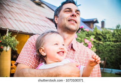 Funny Portrait Of A Happy Father Who Holds His Laughing One Year Old Daughter - Sweet Little Baby Girl Toddler. Happy Fathers Day Concept Photo - Face Closeup In The Garden In Front Of A House