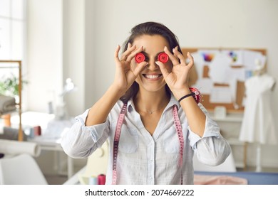 Funny portrait of happy professional dressmaker, seamstress or tailor at work in modern atelier. Positive cheerful young woman with measuring tape around neck covers eyes with two red plastic buttons