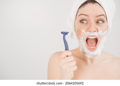 Funny portrait of a woman with shaving foam on her face holding a razor on a white background. The girl removes the mustache and beard