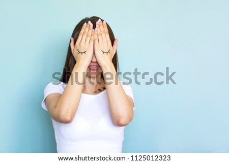 Funny portrait of young brunette woman with eyes painted on hands. Model with closed eyes on blue background. Creative concept of beauty, fun. Copy space.