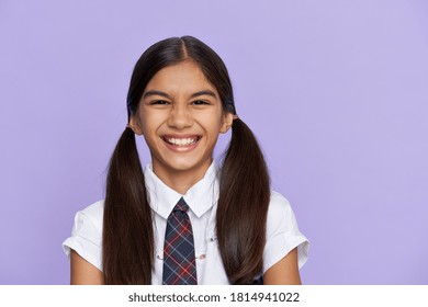 Funny positive indian kid primary school girl with ponytails wearing uniform laughing grimacing looking at camera isolated on violet background. Happy latin child student close up headshot portrait.