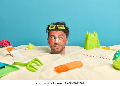 Funny Positive Man Has Head Stuck In Sand, Wears Goggles, Sunburnt Red Skin, Relaxes During Summer Weekend, Enjoys Good Day, Sunbathing, Looks Aside With Interest, Spends Time At Sandy Beach